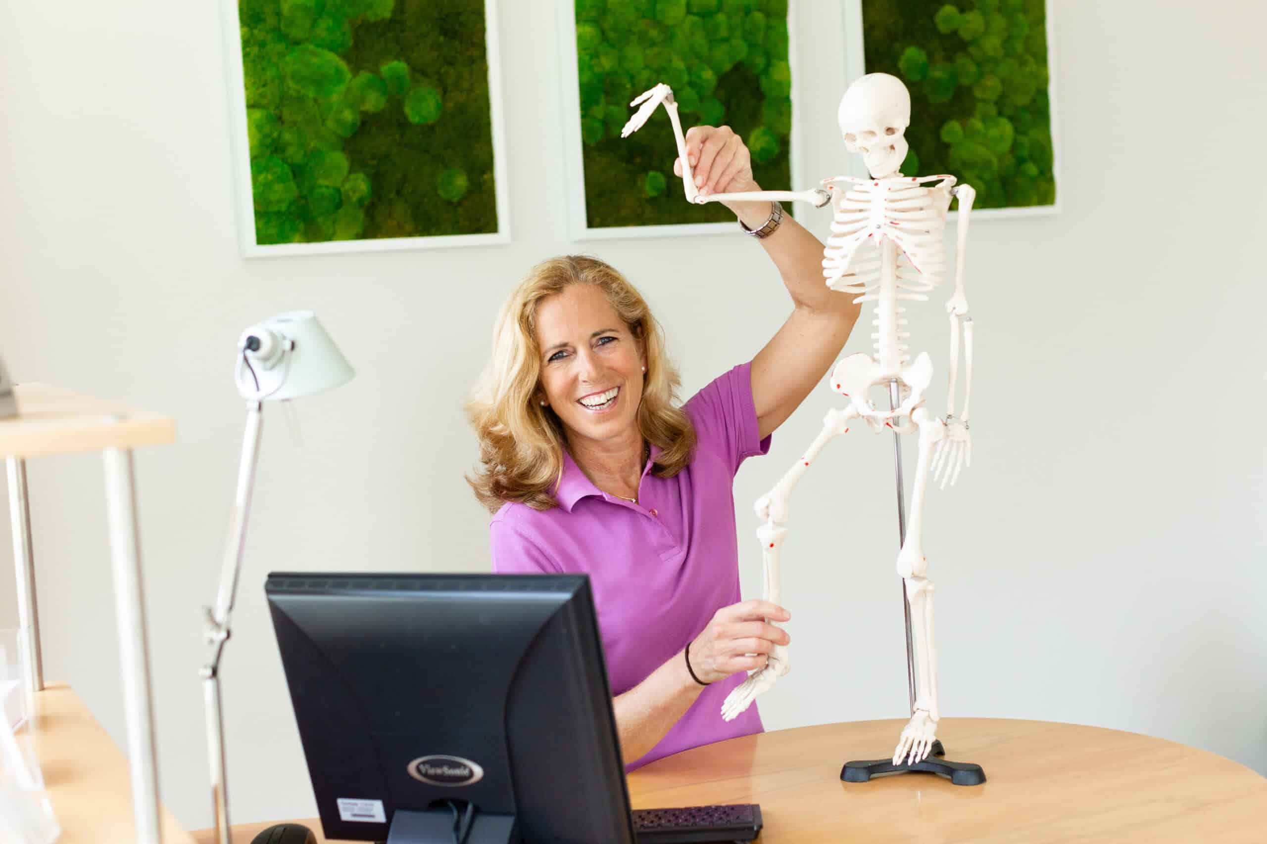 Sigrid Melzer, Frau mit blondem Haar und lila Polo, sitzt lächelnd an ihrem Bürotisch mit einem Modell-Skelett in ihren Händen